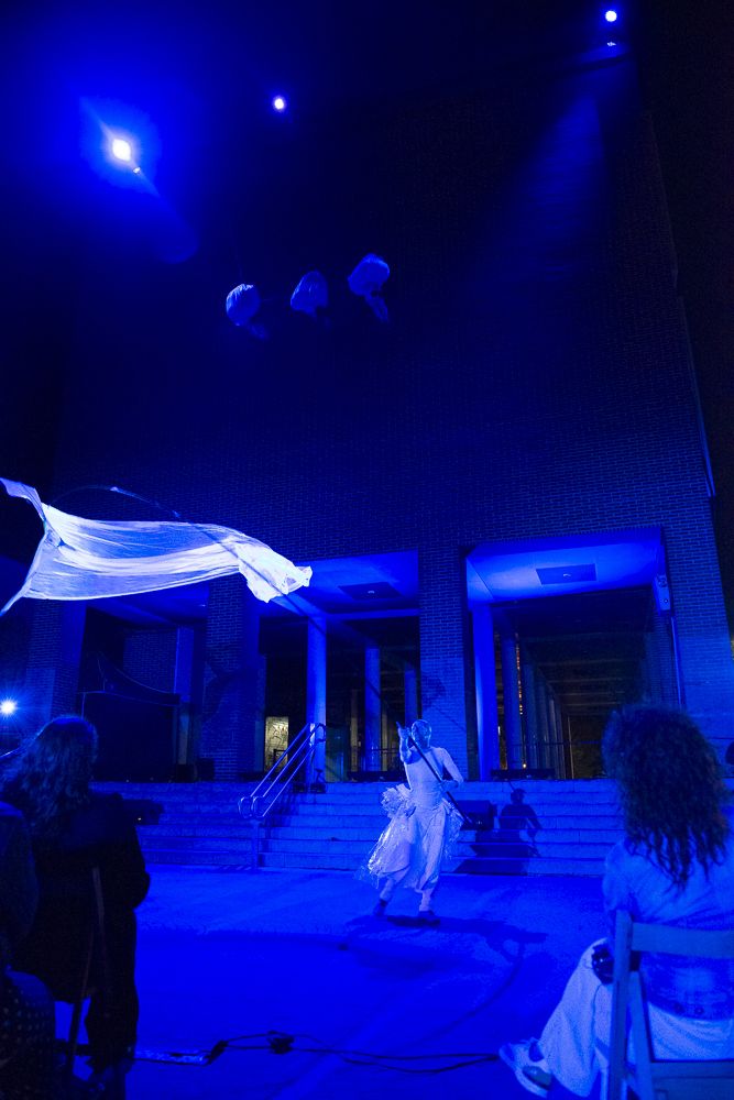 Performer in white guiding a flying fabric bird on a pole in blue light in front of a brick building, Eco Flight tour