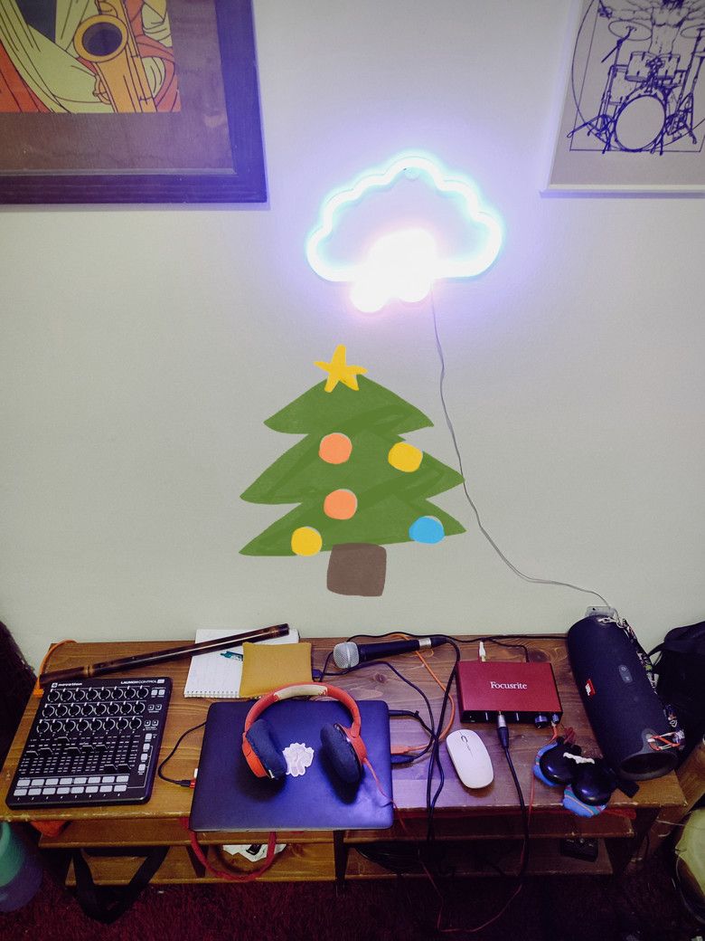 Luca Aza's home studio desk with a Novation Launch Control, Focusrite audio interface, laptop, headphones, microphone and flute under a neon cloud light and a painted Christmas tree