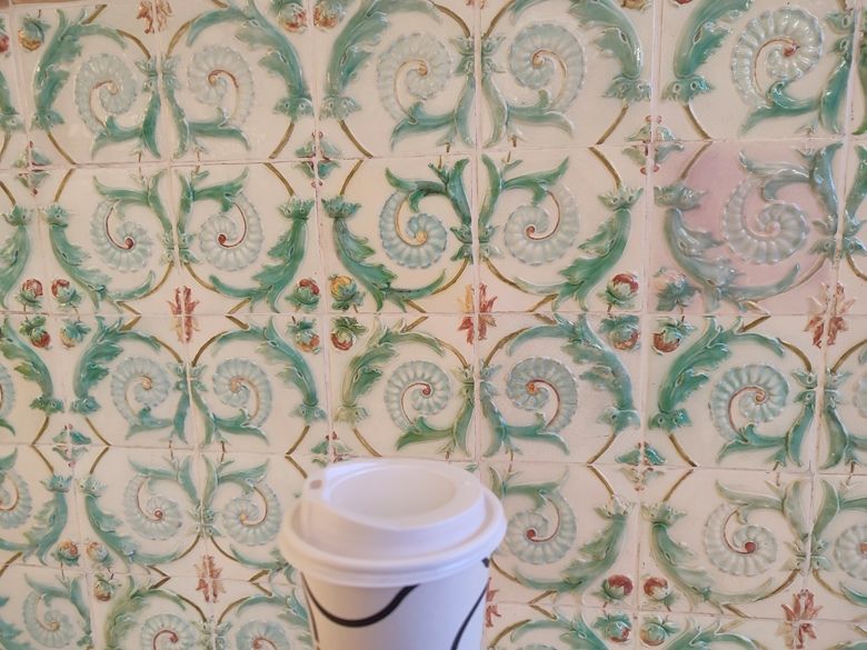 Takeaway coffee cup in front of green and white ornamental ceramic tiles at the University of Pécs art department